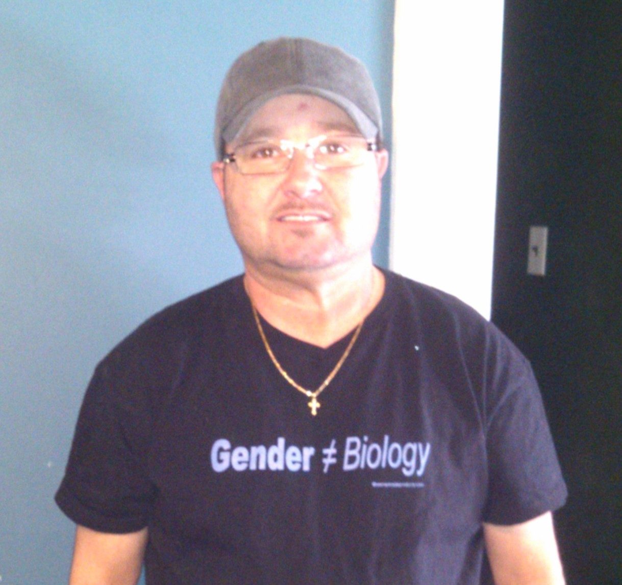 BJ Buchanan, Master Plumber in Newport News, VA and across Hampton Roads, appears in a head-and-shoulders shot with a grey ballcap and black t-shirt that says "Gender ≠ Biology." He stands in front of a blue and white wall.
